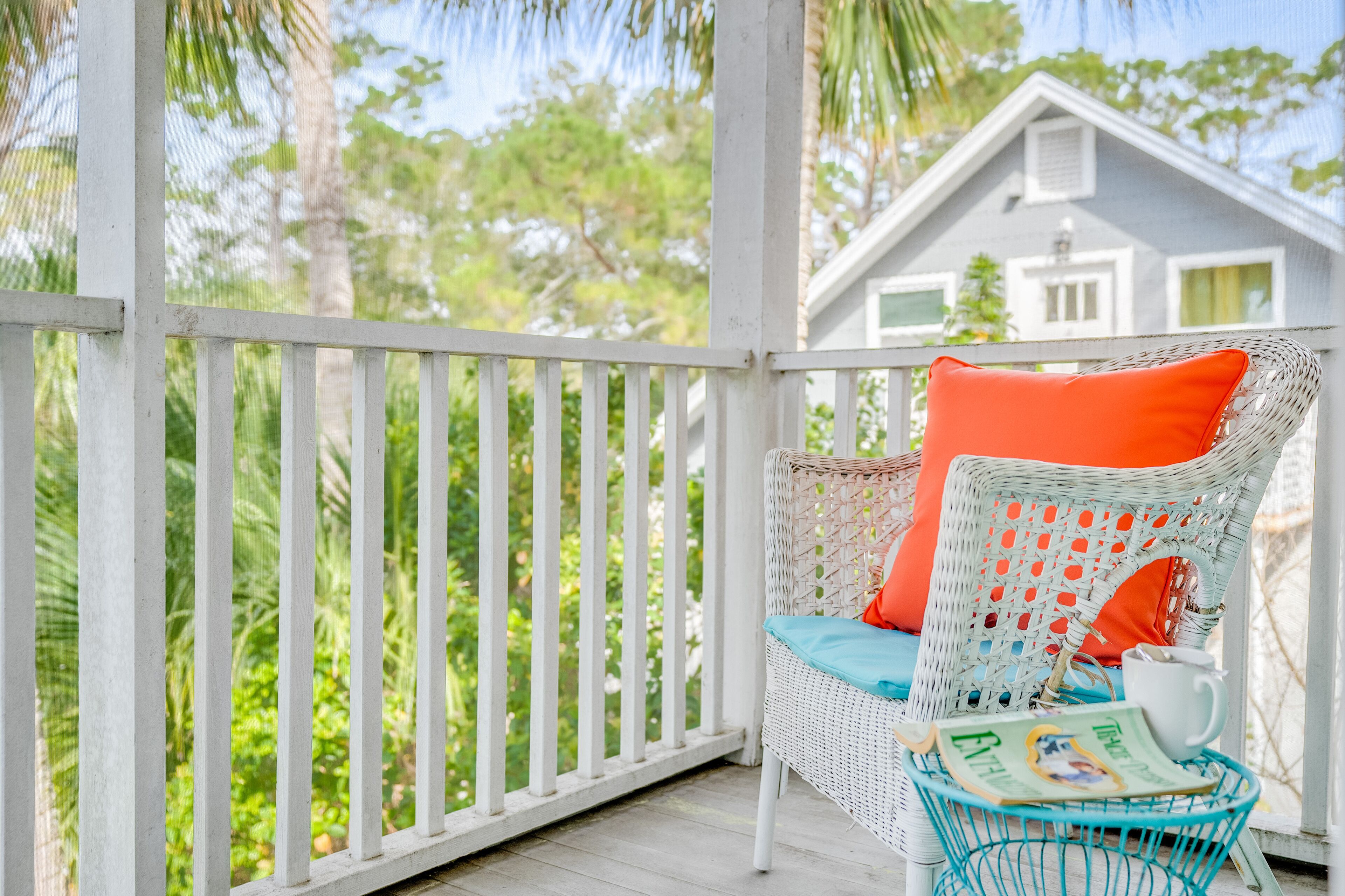 A white wicker chair with an orange cushion on a porch, a teal basket with a magazine, and a white house in the background.