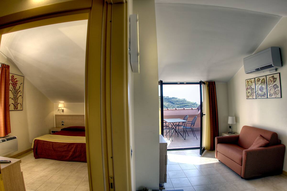 A hotel room with a bed, a sofa, a small table, and a balcony with a table and chairs.
