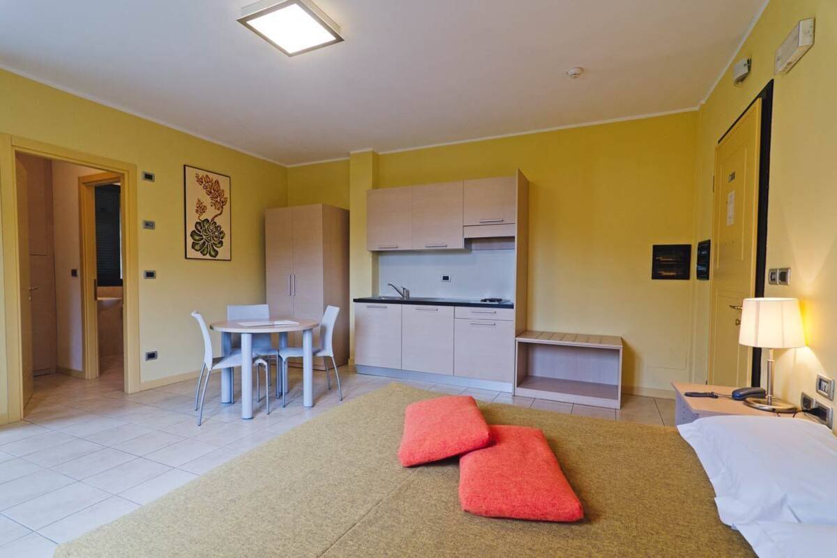 A compact hotel room with a kitchenette, a dining area, and a bed with red cushions.