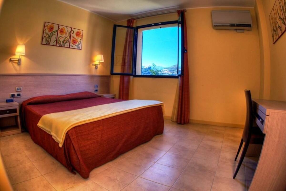 A hotel room with a bed, a desk, a chair, a window with curtains, an air conditioner, and wall-mounted lamps.