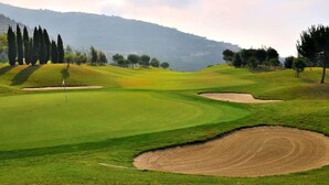 En golfbana med sandbunkrar, en flaggstång och ett kuperat landskap i bakgrunden.
