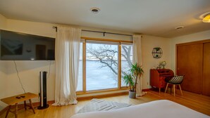 A bedroom with a large window, a bed, a desk with a chair, a TV, and a plant.