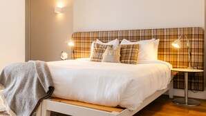 A neatly made bed with a plaid headboard, a bedside lamp, and a small round table with a lamp. - The Saddle and Stable Rooms (Penzance)