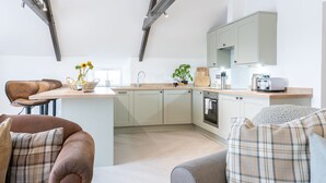A modern kitchen with a central island, built-in cabinets, and a dining area with a brown leather chair. - The Saddle and Stable Rooms (Penzance)