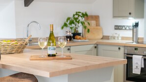 A kitchen counter with a bottle of wine and two glasses, a basket of fruits, and a plant in the background. - The Saddle and Stable Rooms (Penzance)