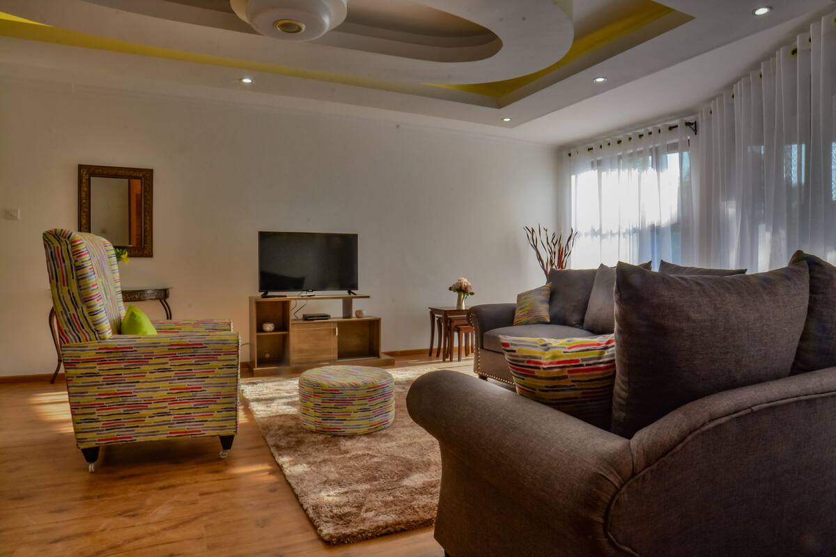 Una sala de estar moderna con sofá, sillón, reposapiés, televisor y un jarrón decorativo.