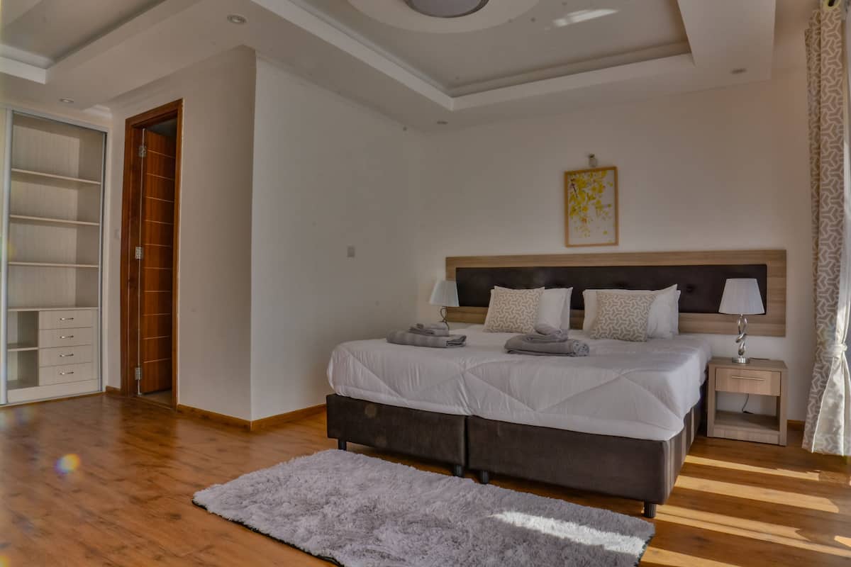 Un dormitorio con una cama grande, suelo de madera, un tapete gris, una mesita de noche con lámpara y un armario.