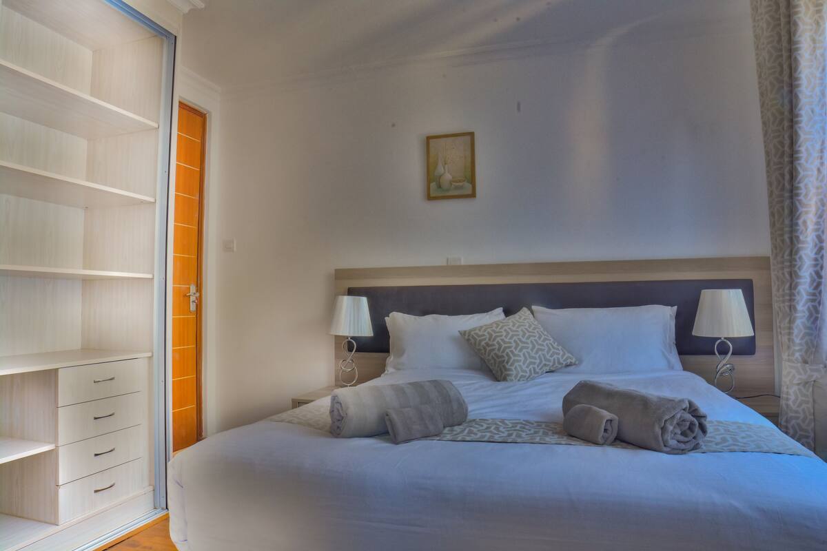 Una cama bien hecha con almohadas y una colcha, una mesita de noche con una lámpara, un armario con las puertas abiertas y un cuadro en la pared.