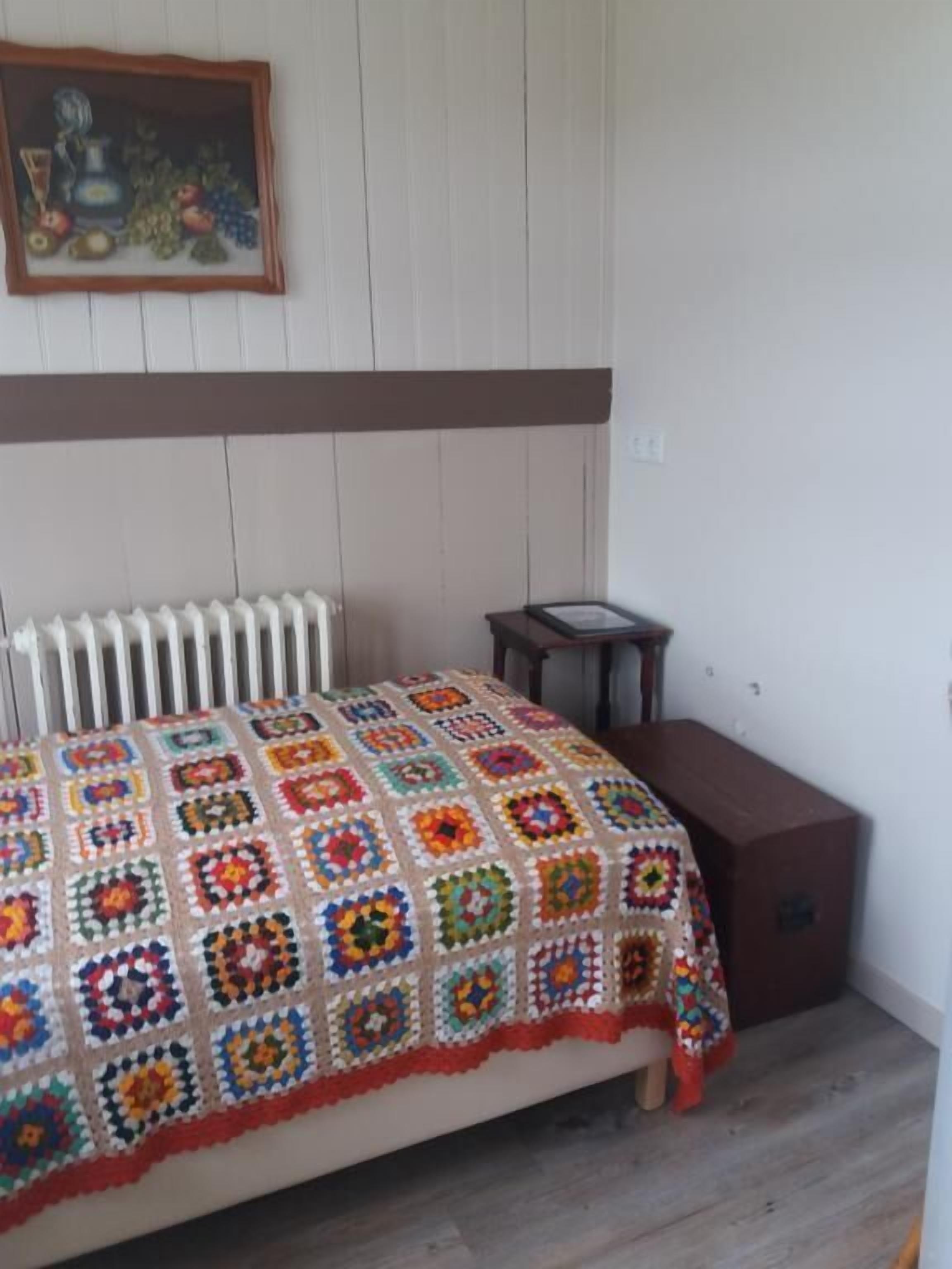 Ein Schlafzimmer mit einer bunten Häkeldecke, einem Heizkörper, einem kleinen Tisch und einem Schrank.