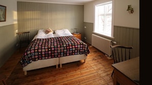 Ein Schlafzimmer mit Holzboden, einem Bett mit farbenfroher Steppdecke, einem Nachttisch mit Lampe und einem Fenster mit Blick auf Bäume.