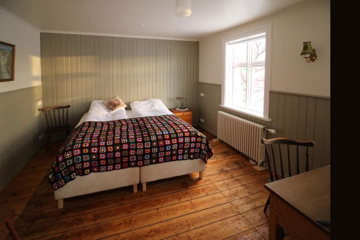 A bedroom with a wooden floor, a bed with a colorful quilt, a nightstand with a lamp, and a window with a view of trees.
