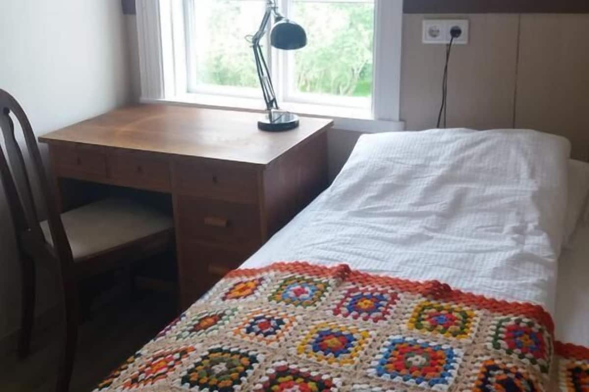 A room with a wooden desk, a chair, a bed with a white sheet, and a colorful crocheted blanket.