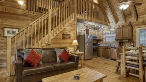Interior de una cabaña de madera con una escalera, un sofá de cuero, una mesa de madera y una zona de cocina.