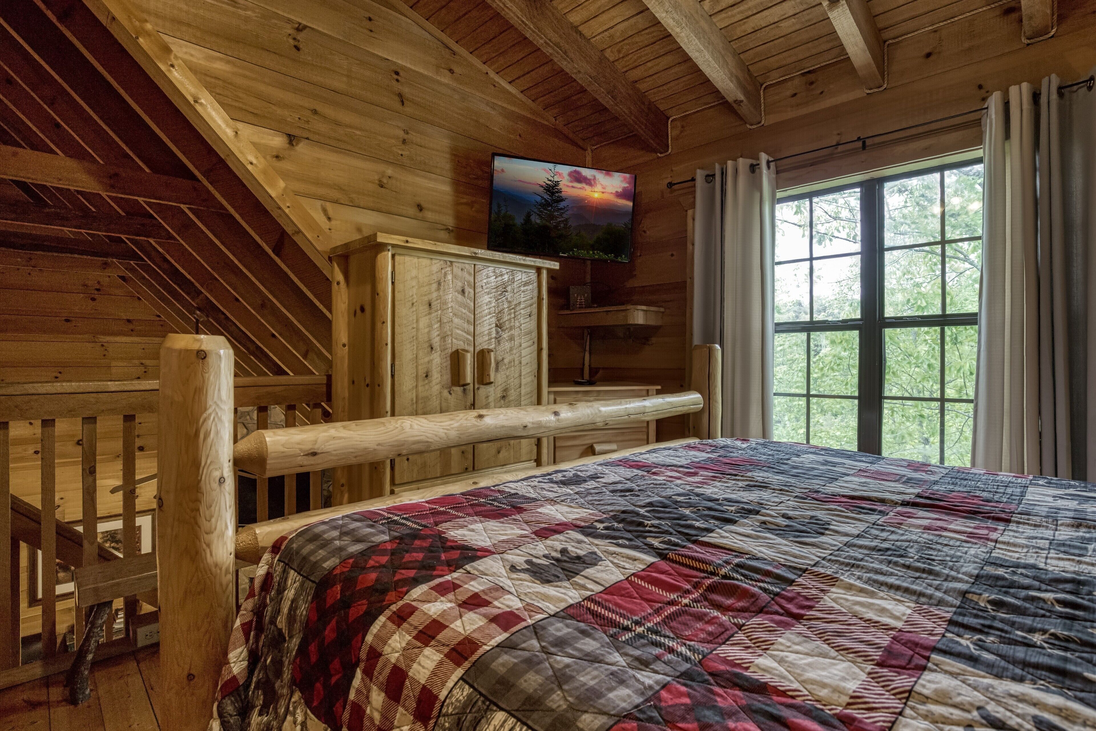 A wooden cabin room with a bed, a television on the wall, and a window with curtains.