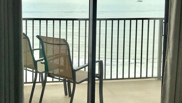 A room with a large sliding glass door leading to a balcony overlooking the sea. There is a chair on the balcony and a bed with a green quilt in the room.