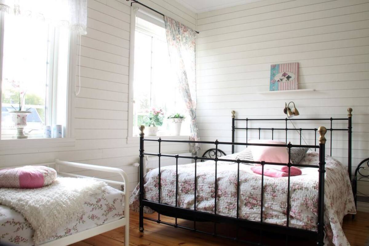 A bedroom with a black metal bed frame, a white bed with floral bedding, a window with curtains, and a small shelf with a picture.