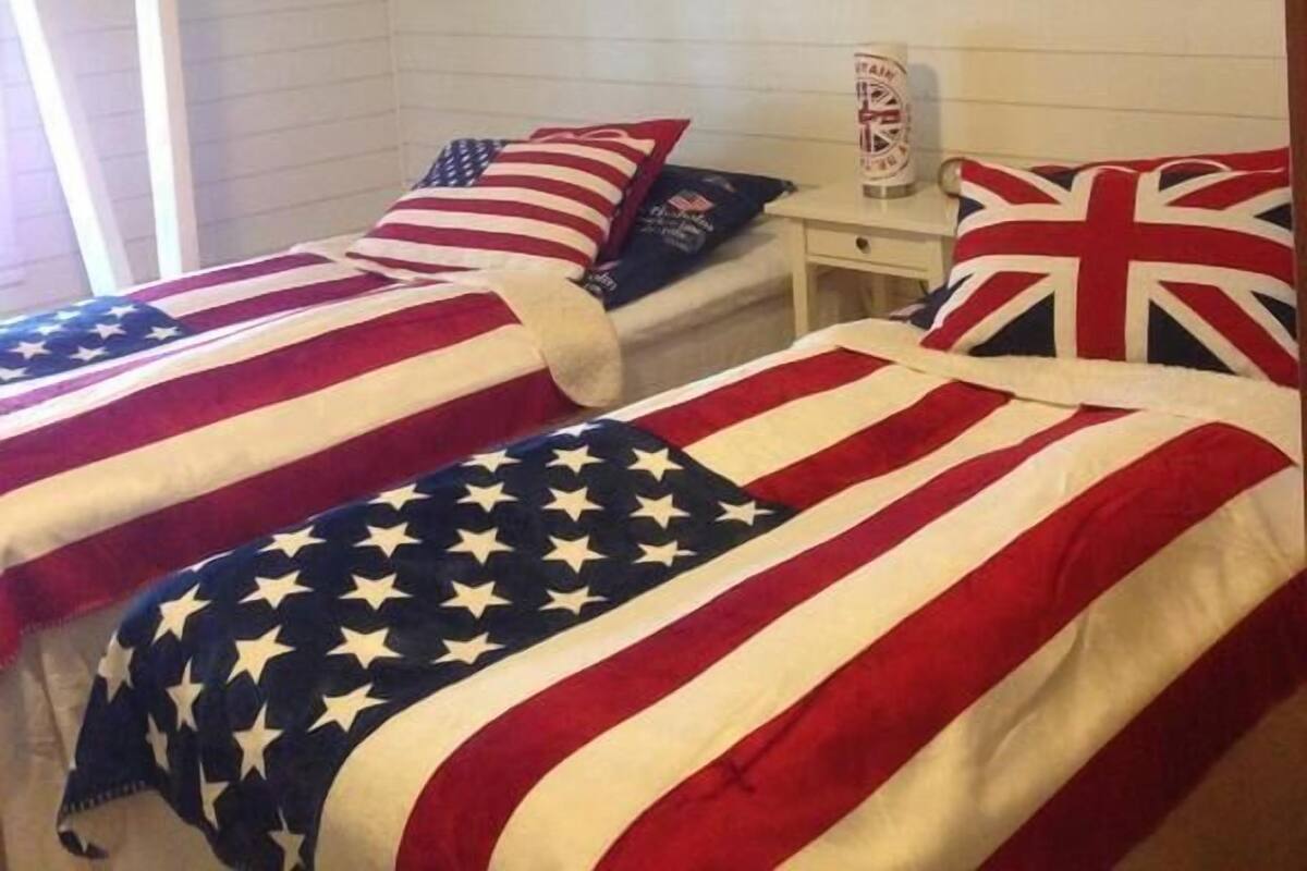 A room with two beds, each covered with an American flag blanket. There is a small table with a clock and a framed picture of the Union Jack on the wall.