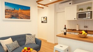 En moderne stue med en blå sofa, et træbord med frugt, og et køkkenområde med hvide barstole og en mikrobølgeovn.