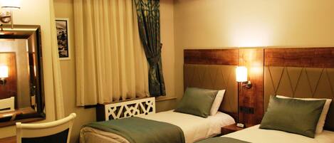 Habitación de hotel con dos camas individuales, un escritorio con silla, un espejo, una mesita de noche con lámpara y una ventana con cortinas.