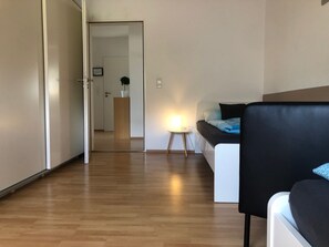 Moderni huone, jossa on puulattia, musta sohva, pieni yöpöytä lampun kanssa ja ovi, joka johtaa toiseen huoneeseen.