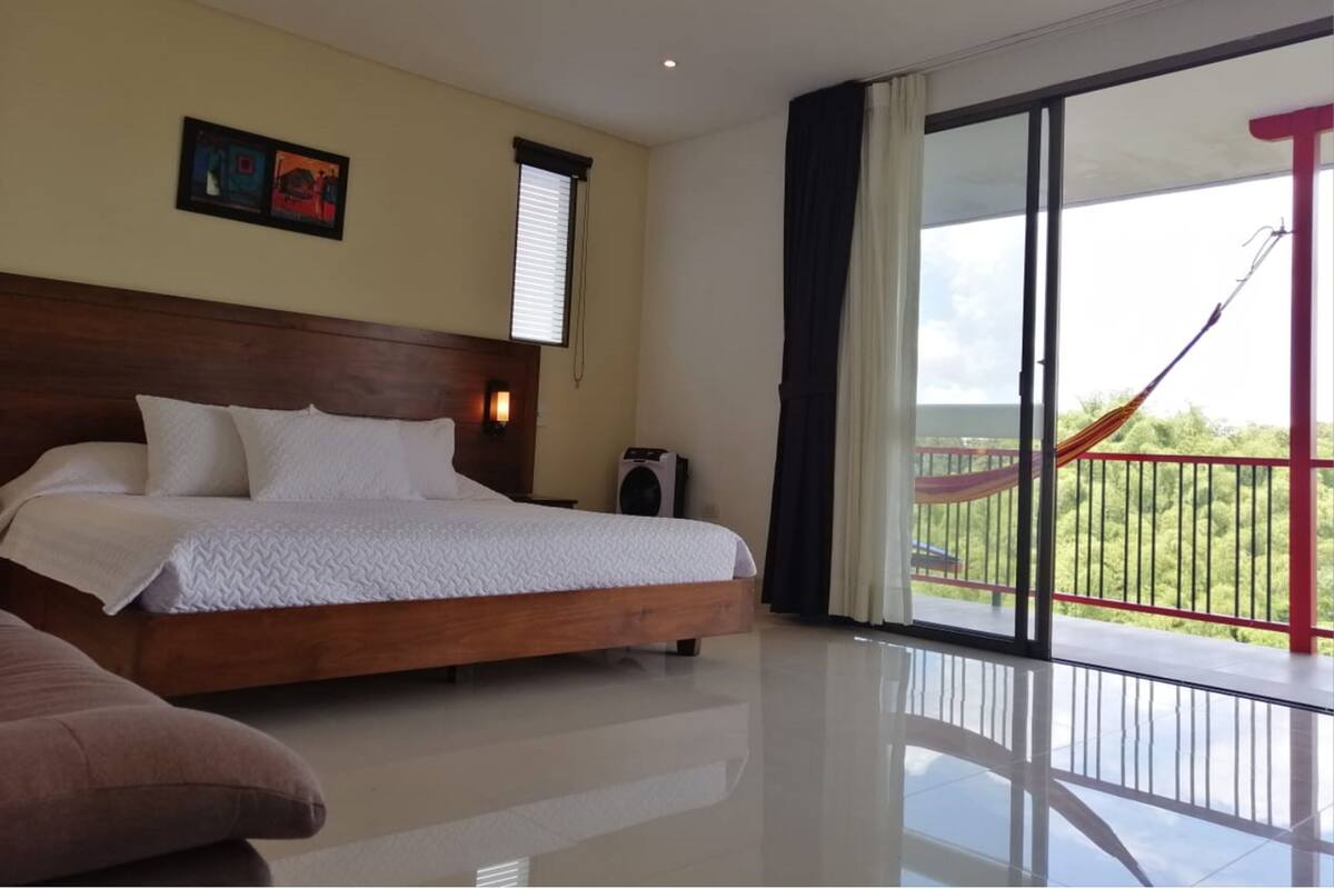 Une chambre d’hôtel avec un grand lit, une petite table, une chaise, une fenêtre avec des stores, une lampe fixée au mur et un balcon avec un hamac.