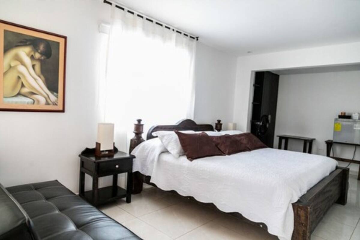 Une chambre à coucher avec un lit, une table de chevet, une lampe, un tableau au mur et une fenêtre avec des rideaux.