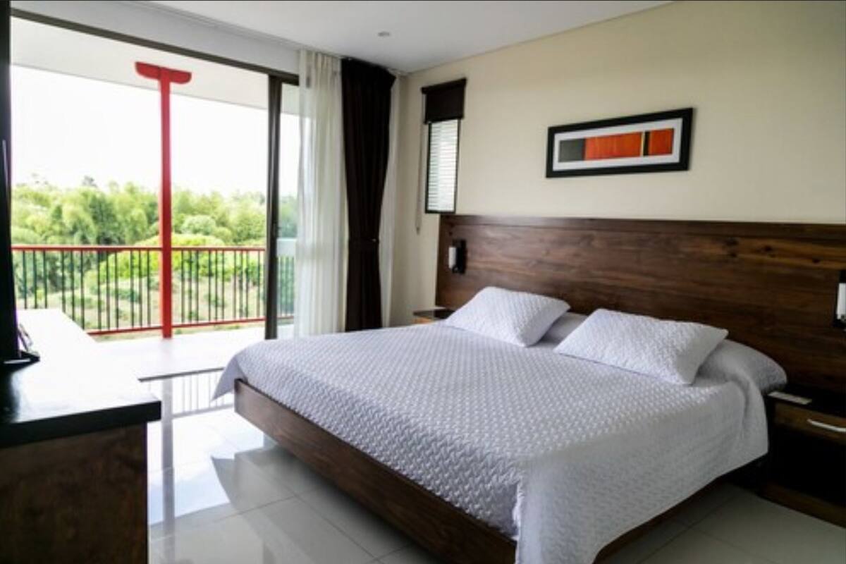 Une chambre avec un grand lit, une tête de lit en bois, une table de chevet, un bureau avec une chaise et un balcon doté d’une rambarde rouge.