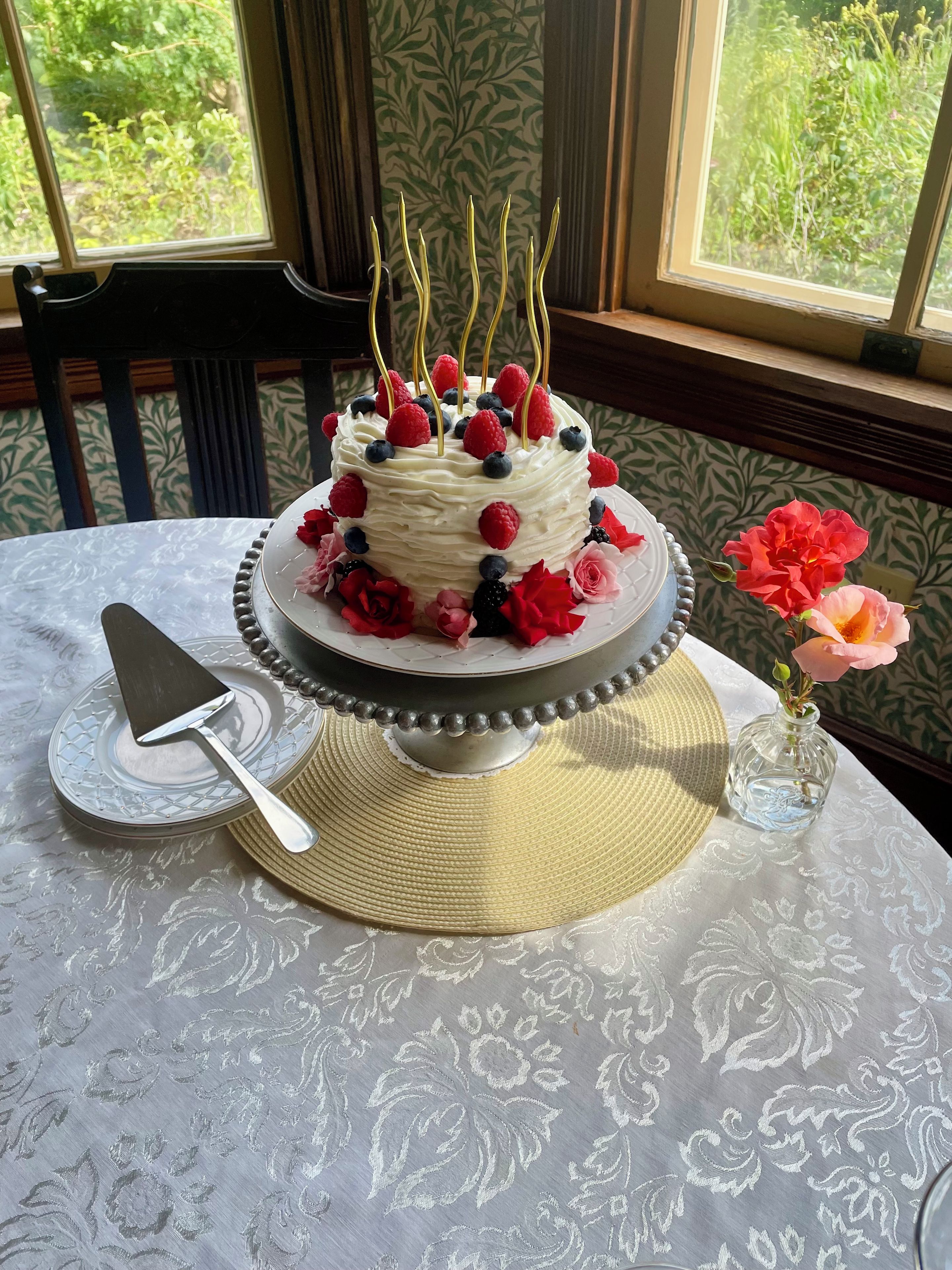 A cake with fruit on a stand, a cake server, and a vase with flowers on a table.