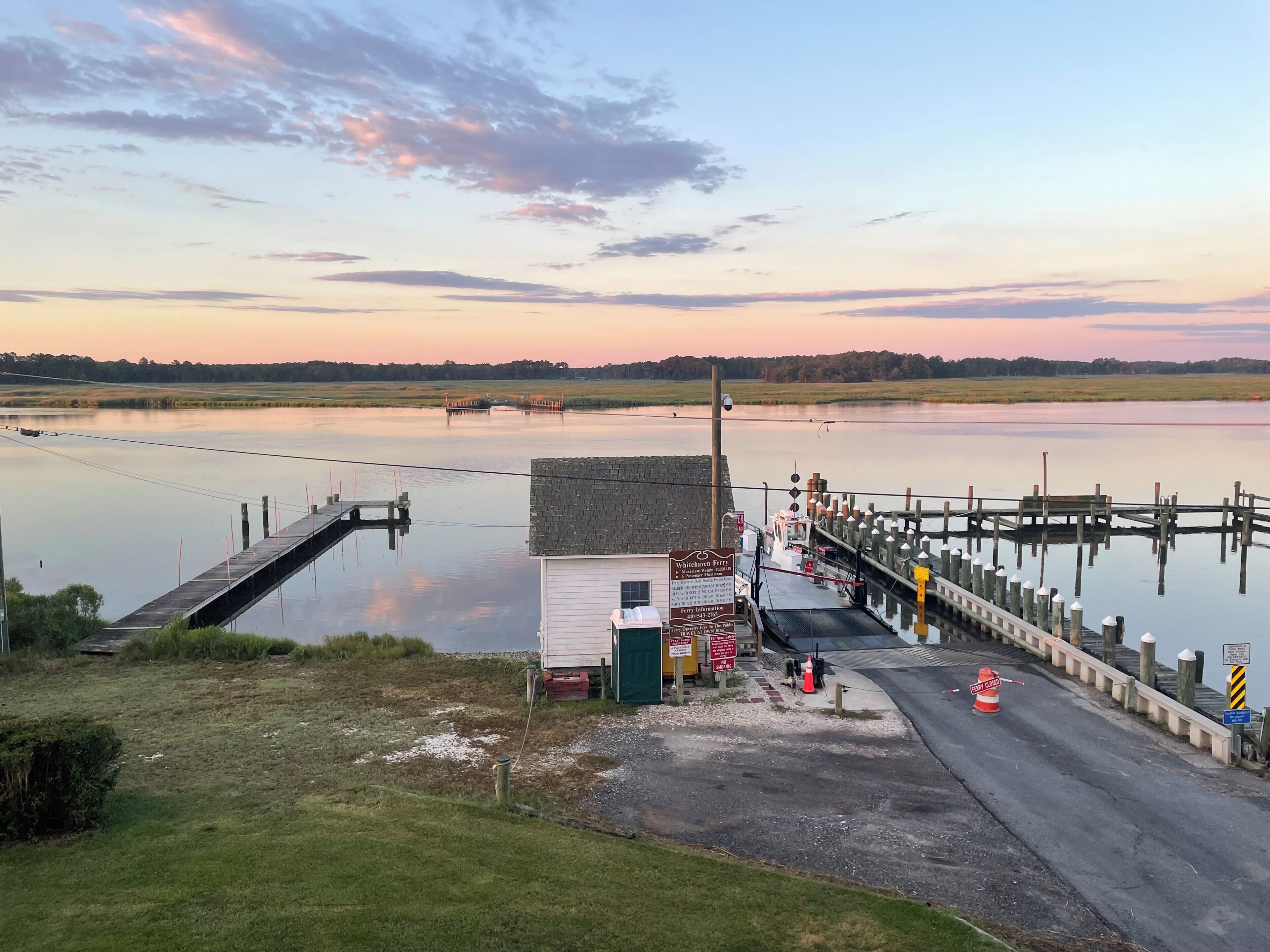Ein Fährterminal mit einer Anlegestelle, die in ruhiges Wasser ragt, umgeben von Feldern und einem klaren Himmel.
