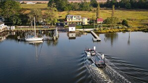 Un río con un muelle, un velero y un barco a motor. Se ven edificios y árboles al fondo.