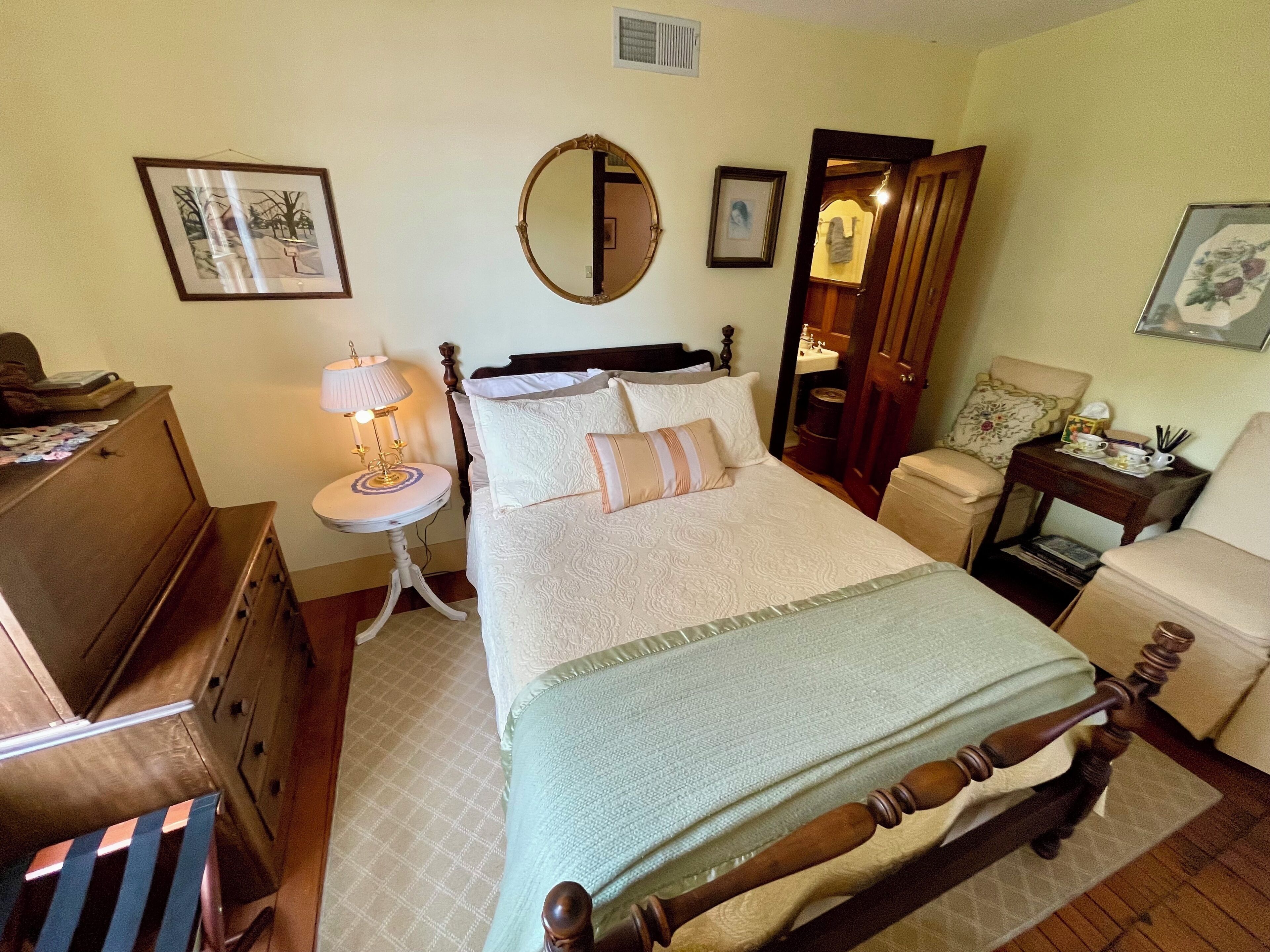 Un dormitorio con cama, cómoda, mesita de noche, silla y un baño visible a través de una puerta abierta.
