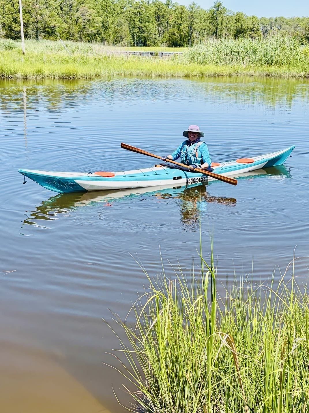 A person in a canoe on a calm body of water with grassy banks and trees in the background.