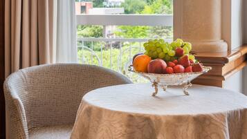 Una habitación con vistas a una colina verde, una silla de mimbre, una mesa con frutero y una ventana con cortinas.
