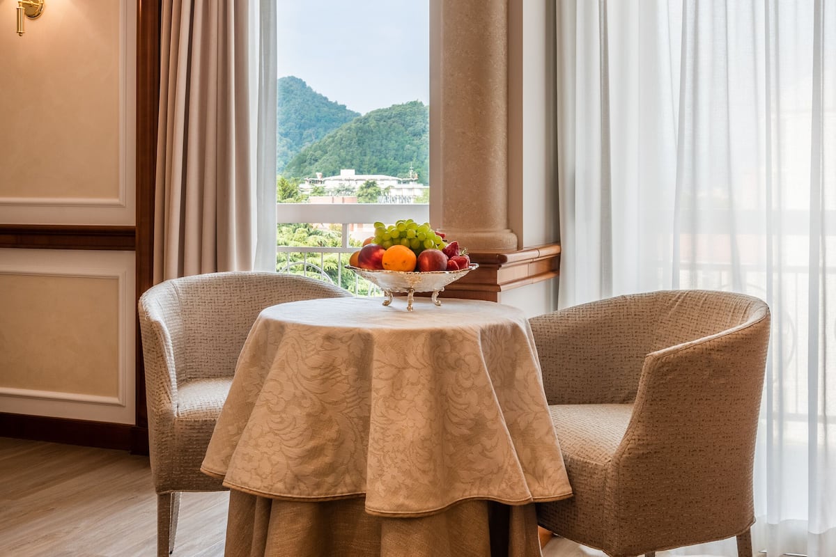 A dining area with a round table, two chairs, a fruit bowl, and a view of greenery through large windows.
