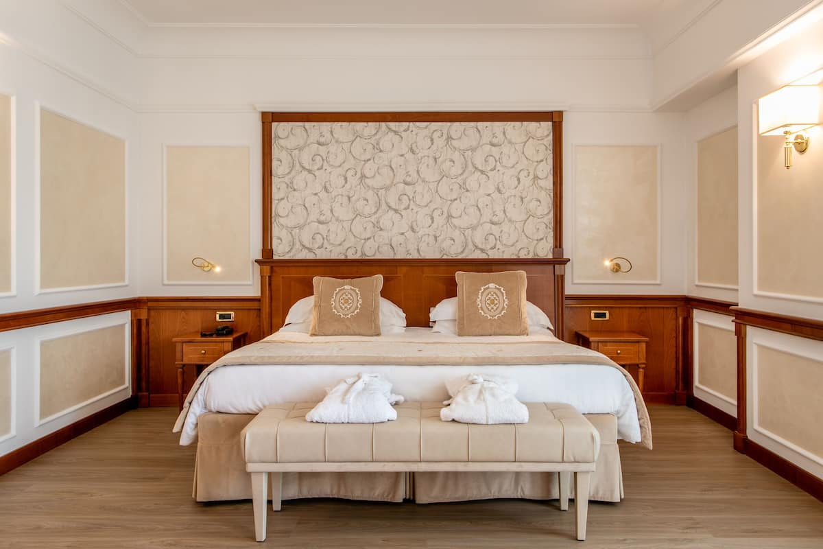 A hotel room with a large bed, two bedside tables, a bench with towels, and a decorative headboard.