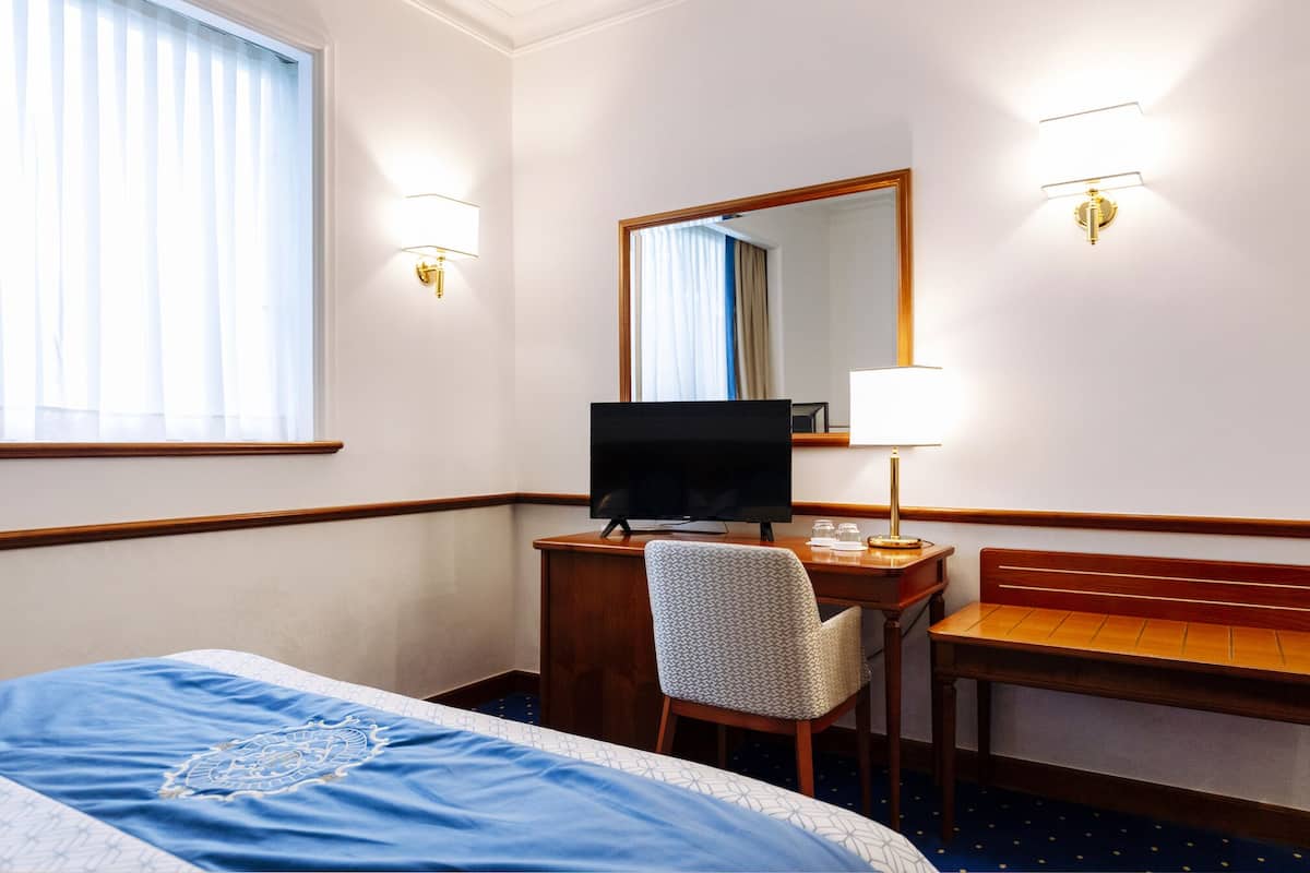 A hotel room with a bed, a desk with a computer, a chair, a desk lamp, and a mirror.