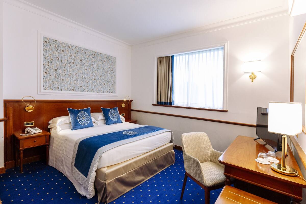 A hotel room with a bed, bedside table, chair, desk, lamp, and a large window with curtains.