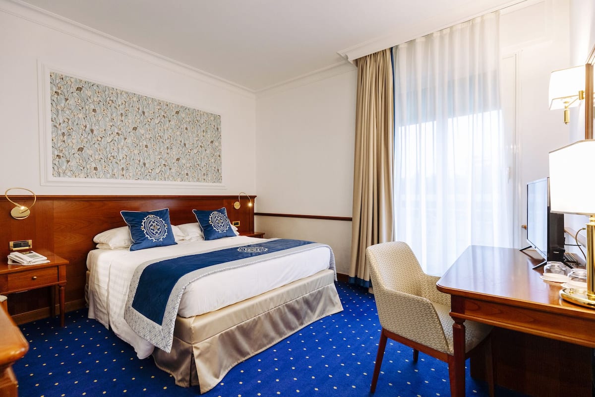 A hotel room with a bed, a desk, a chair, a lamp, and a painting on the wall.