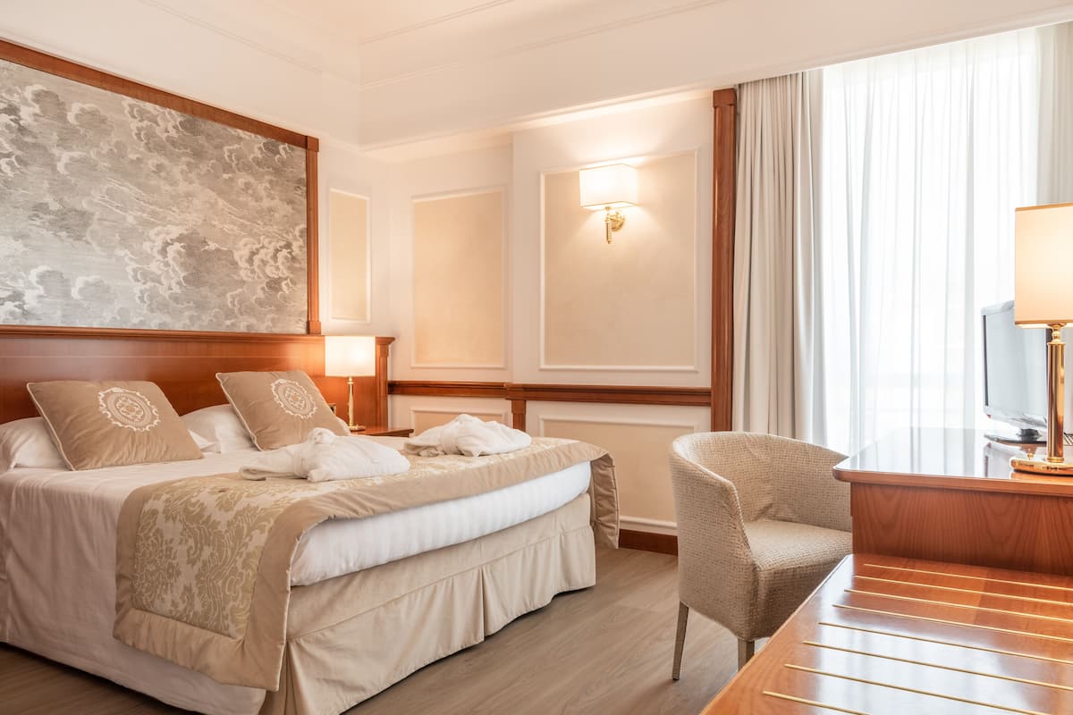 A hotel room with a large bed, a desk with a lamp, a chair, and a television.
