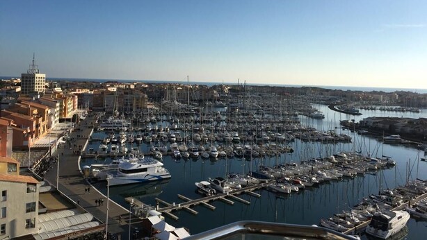 Un puerto deportivo con numerosos barcos amarrados, cielo despejado y edificios a lo largo de la costa.