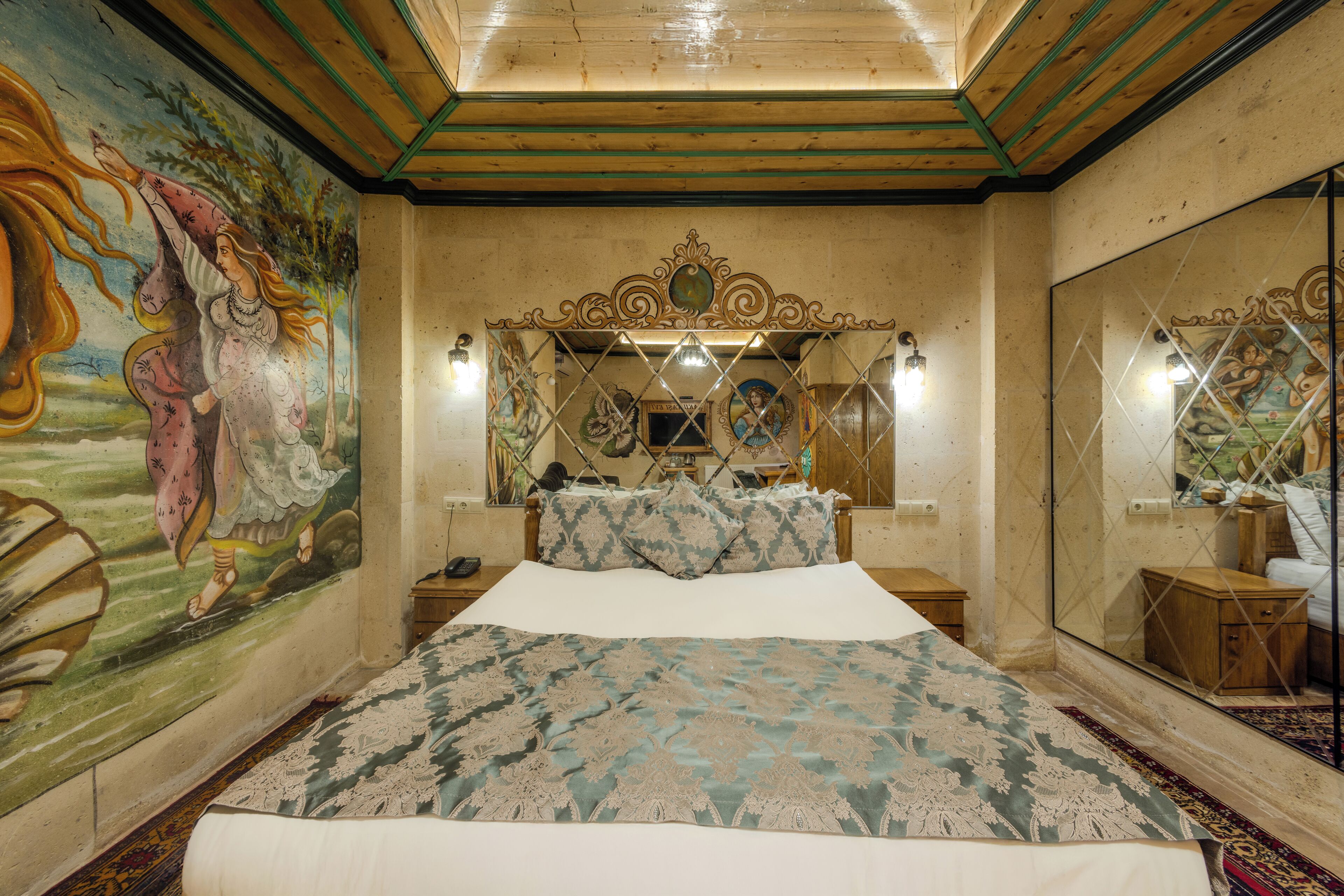 A bedroom with a large bed, a decorative headboard, a mural on the wall, and a mirror reflecting the room.