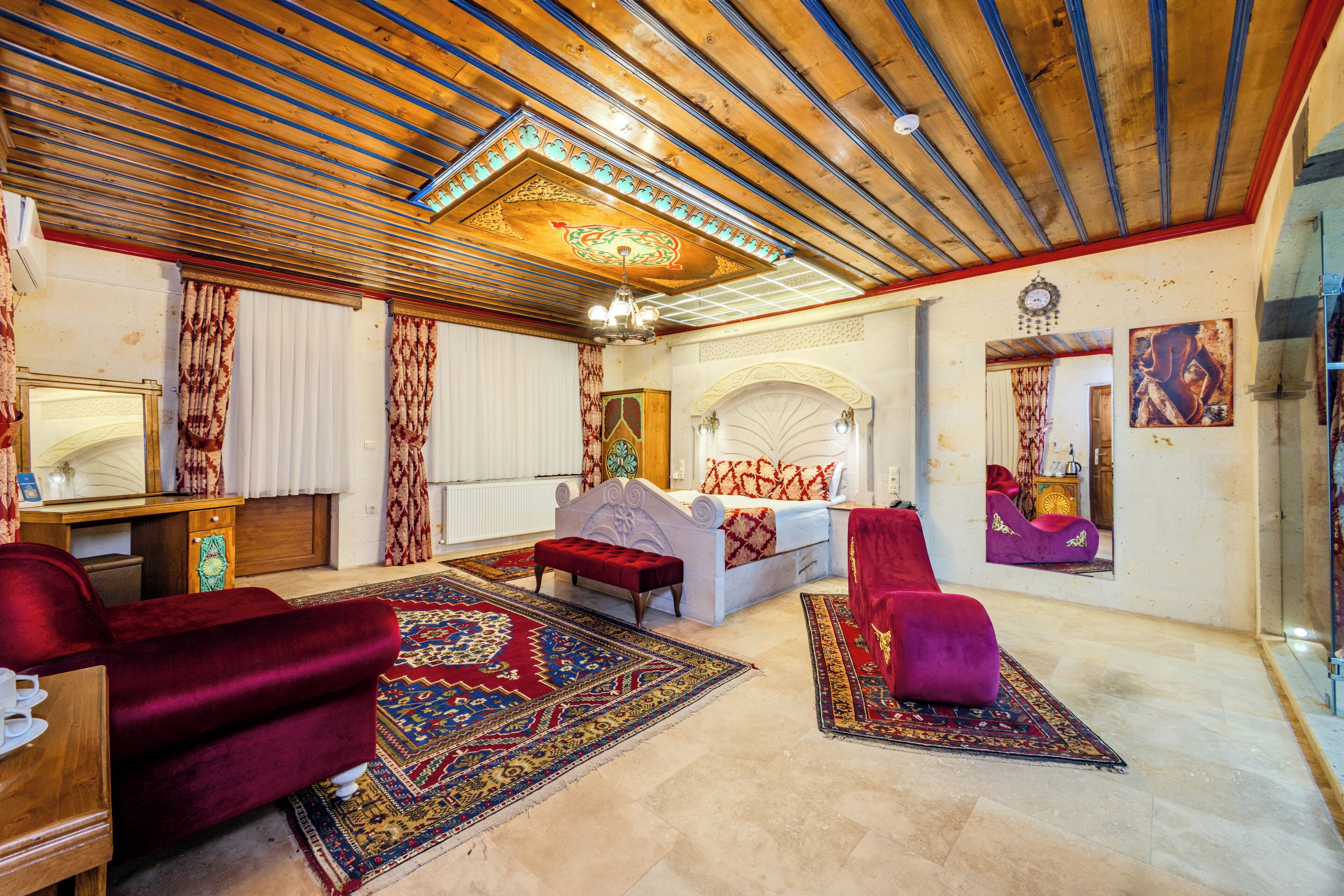 A room with a wooden ceiling, a bed with a red and white patterned bedspread, a red velvet armchair, a purple armchair, and a large ornate rug.