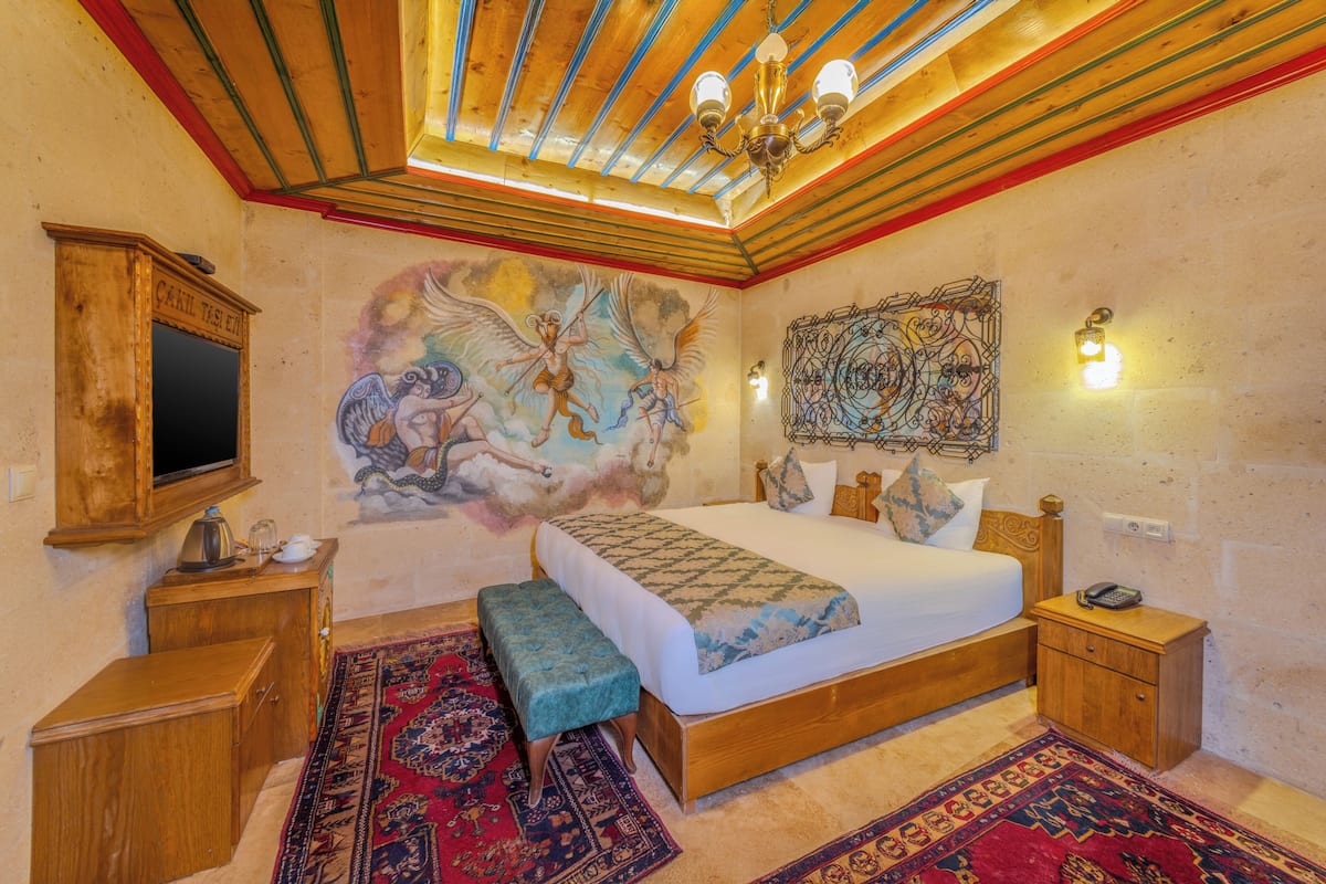 A bedroom with a large bed, a wooden headboard, a bench, a nightstand, a wall-mounted TV, and a ceiling with wooden beams and a chandelier.