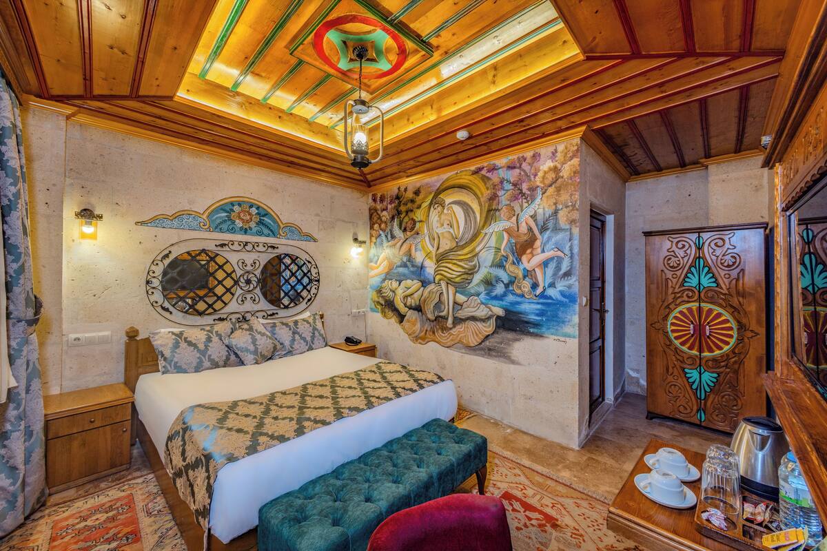 A bedroom with a bed, a bench, a nightstand, a wall mural, a decorative ceiling, and a wooden door.