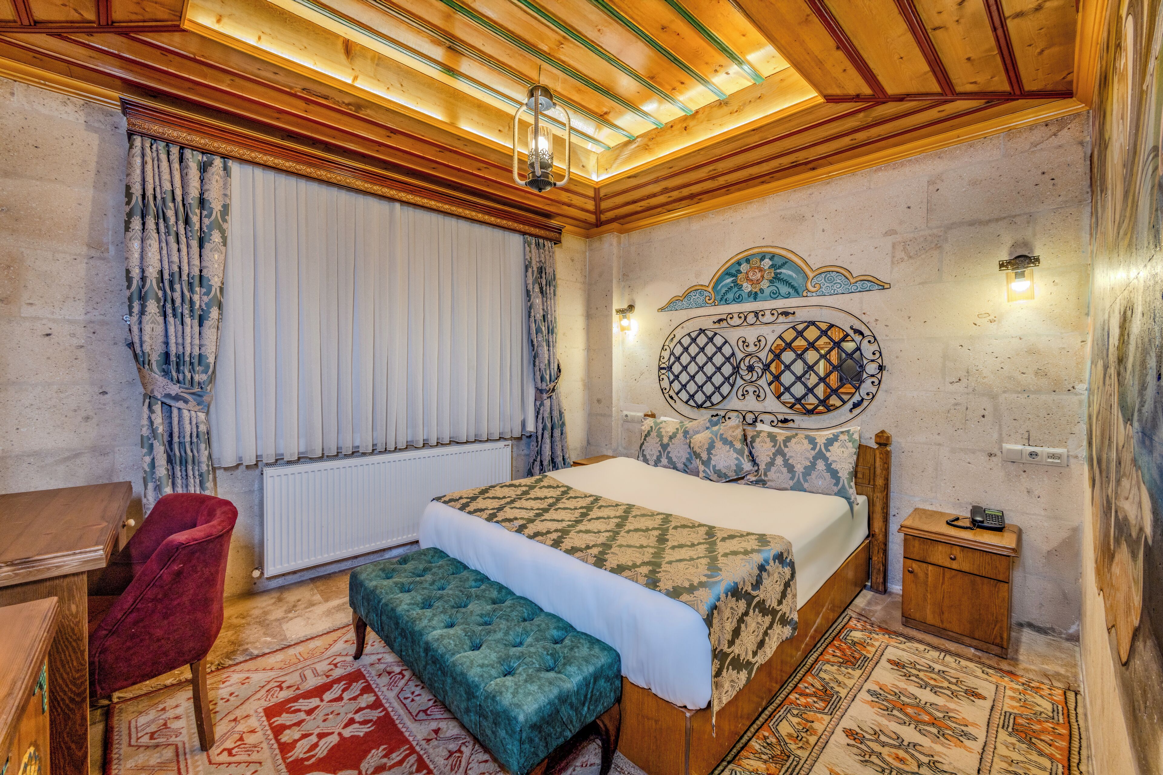 A bedroom with a wooden ceiling, a bed with patterned bedding, a red chair, a bench, a small bedside table, and a wall-mounted light fixture.
