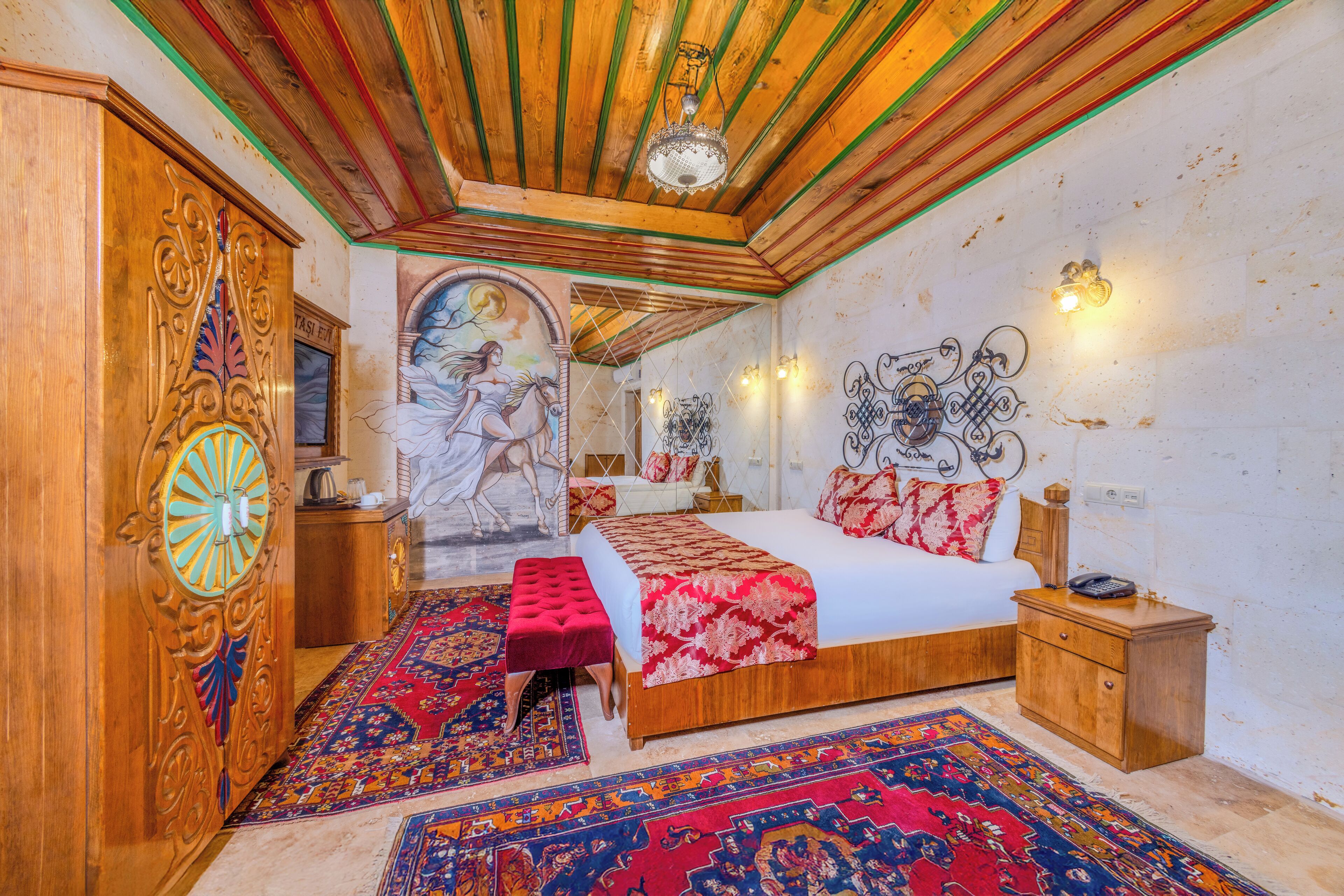 A bedroom with a wooden ceiling, a large bed with red and white bedding, a red ottoman, and a colorful rug.