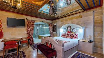 A bedroom with a large bed, a desk, and a bench. There is a TV mounted on the wall, and a chandelier hangs from the ceiling.