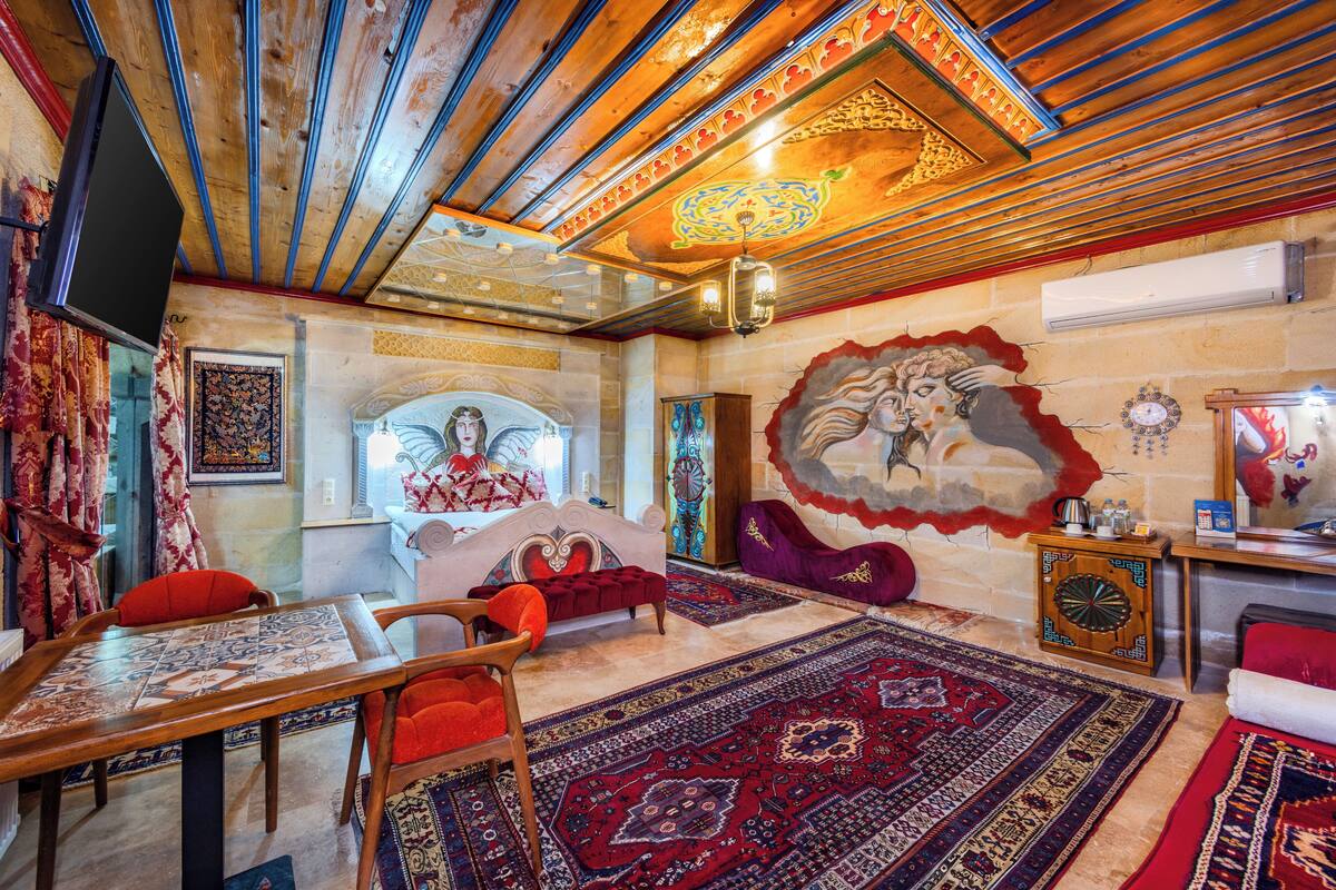A room with a wooden ceiling, a bed with a red headboard, a red sofa, a wooden side table, and a patterned rug.