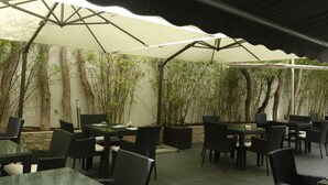 An outdoor patio area with tables and chairs, shaded by large umbrellas, surrounded by bamboo plants.