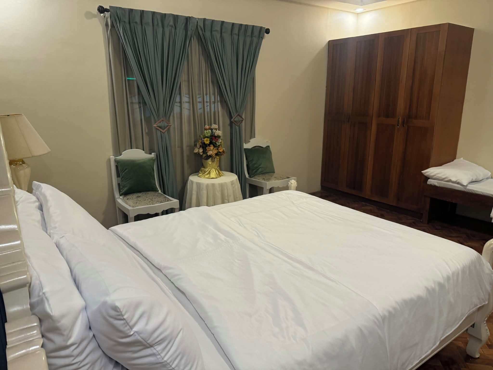 Una habitación de hotel con una cama grande, dos sillas, una mesita con un florero y un armario.
